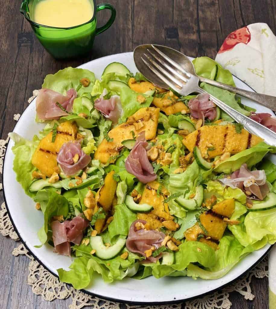 Plate of grilled melon and prosciutto salad with silverware and a small pitcher of dressing.