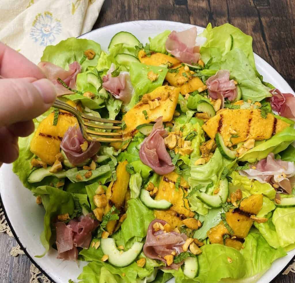 A hand using a fork to stab a piece of cantaloupe from a plate of salad.