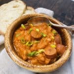 Brown bowl of soup with beans and hotdogs alongside sliced bread.