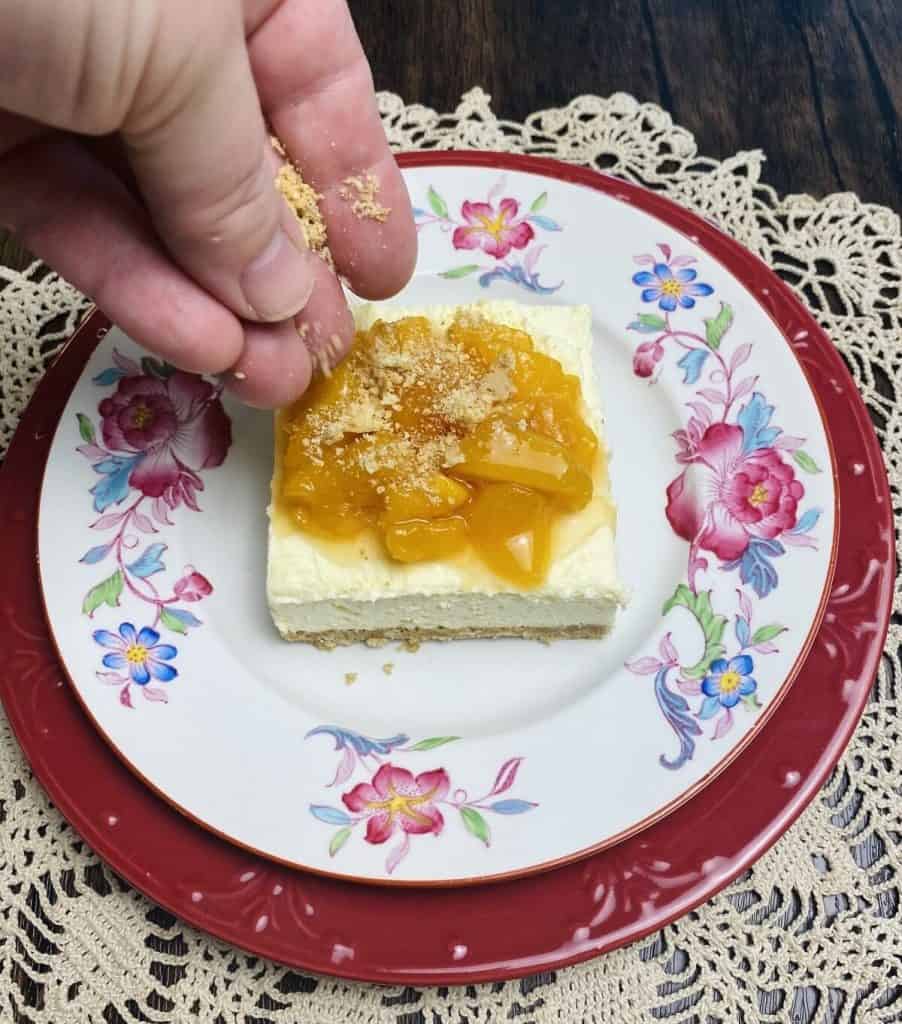 A hand sprinkling graham crumbs onto a piece of peaches and cream dessert.