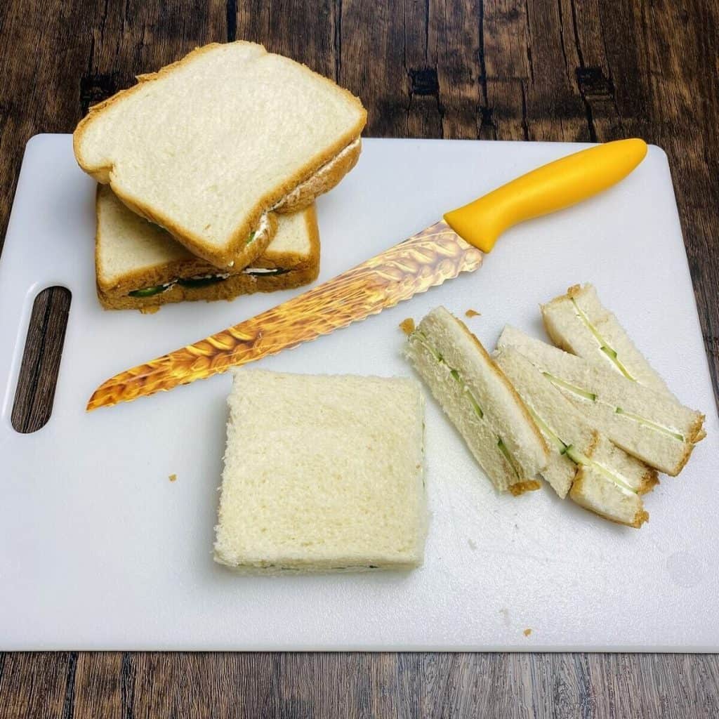 A cutting board with full sized cucumber sandwiches and one has the crusts cut off.