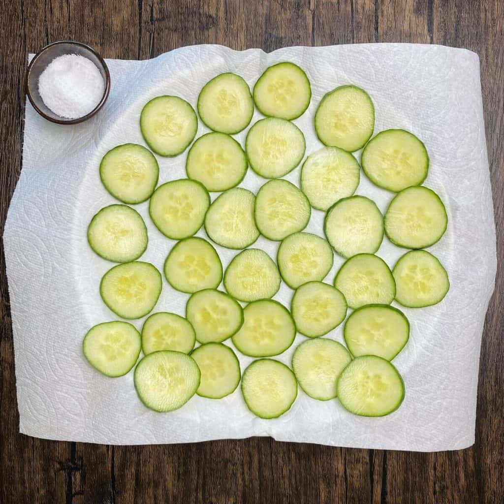 A plate with sliced cucumbers on a paper towel and a small bowl of salt.