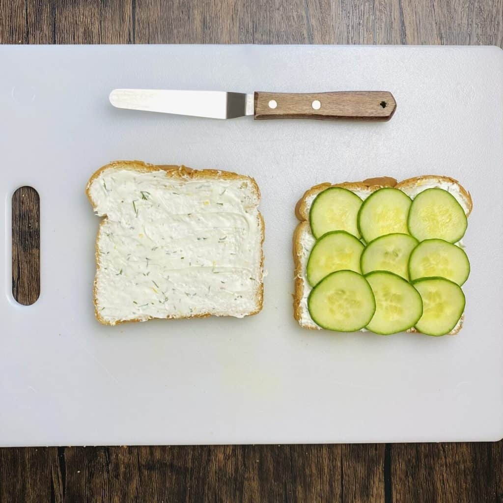 Two slices of bread spread with cream cheese and one side also has sliced cucumber on it.