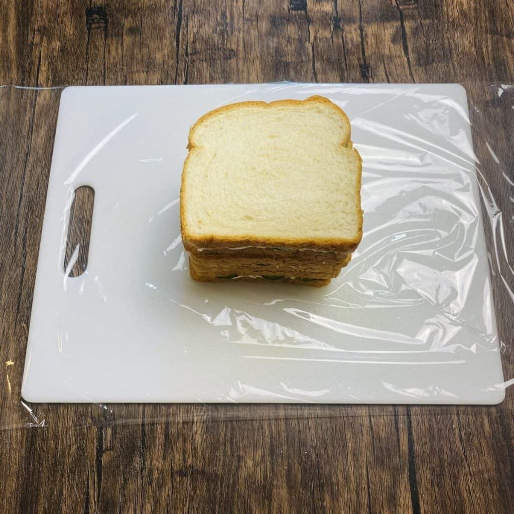 Three whole traditional cucumber sandwiches stacked on a piece of plastic wrap.