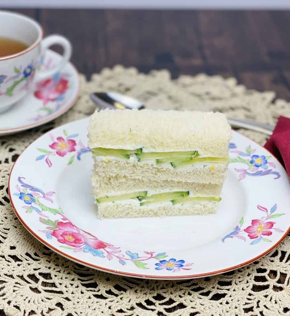 Two traditional cucumber sandwiches stacked on each other on a pretty plate with a cup of tea.