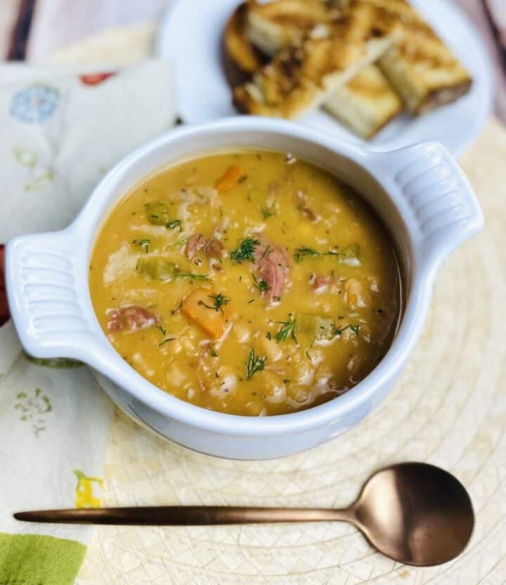 White bowl of ham and beans with grilled bread and a copper spoon.