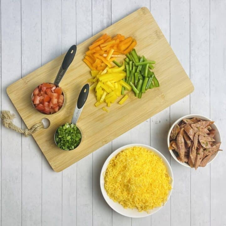 A cutting board topped with chopped vegetables alongside bowls of cheese and sausage.