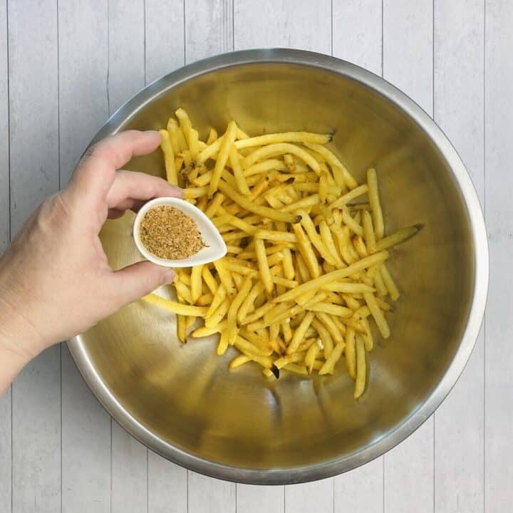 A large bowl of french fries with a hand holding a small bowl of seasoning.