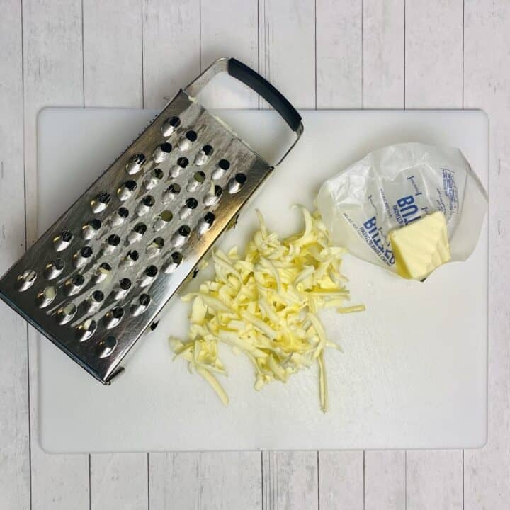 A cutting board with a cheese grater, butter, and shreds of grated butter.