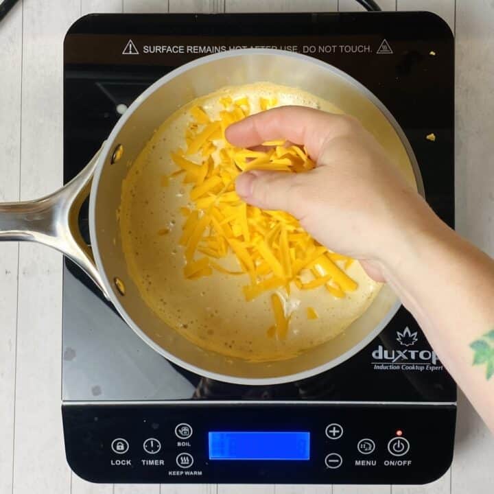 A hand adding shredded cheese to cheese sauce on a burner.