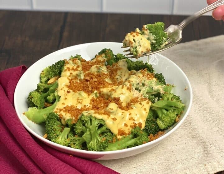 A fork holding up a bite of cheesy broccoli over a bowl of broccoli.