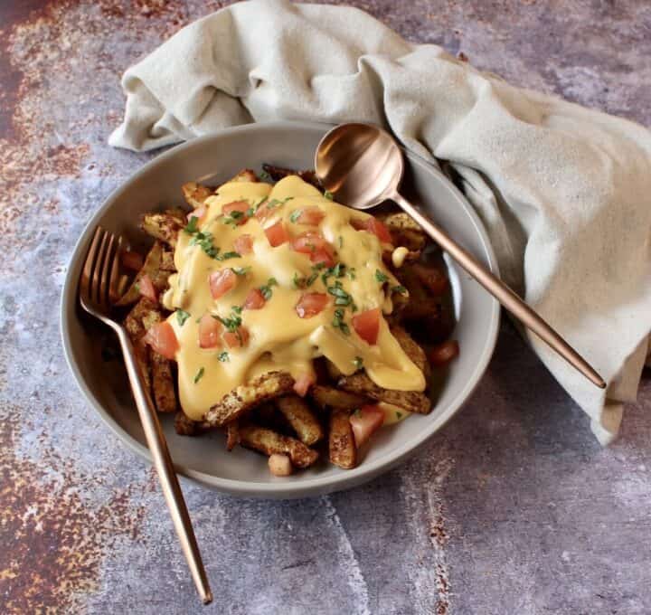 A dish of fries covered in cheese sauce no flour and topped with tomatoes and cilantro with silverware.