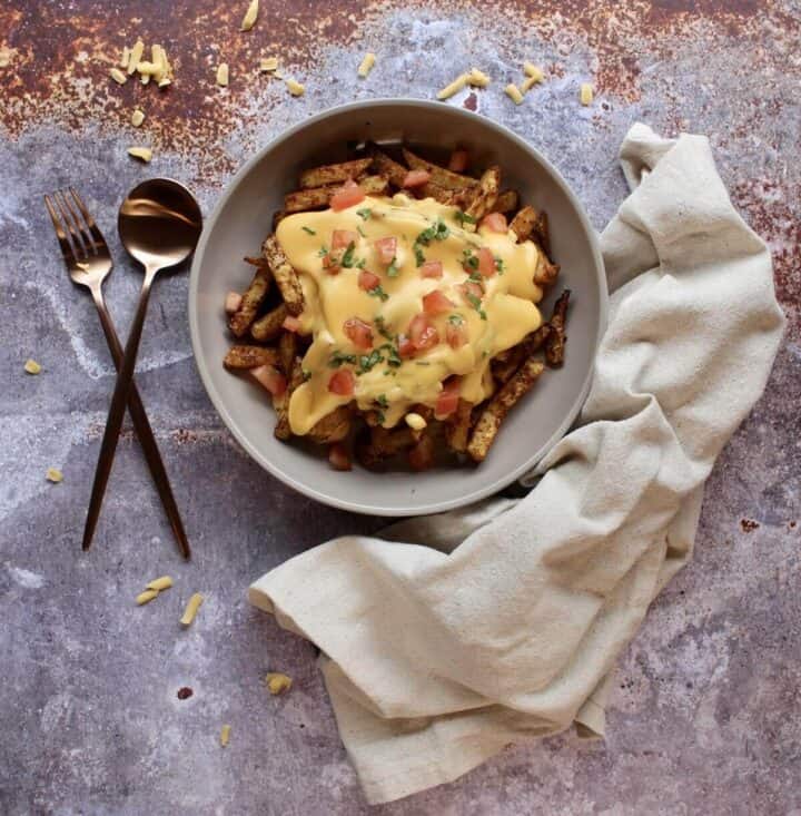 A bowl of fries covered in cheese sauce no flour and topped with garnishes.