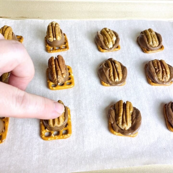 A finger pushing a pecan into a rolo candy on a pretzel.