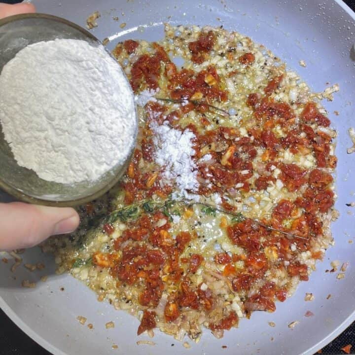 A hand holding a bowl of flour sprinkling into a skillet with recipe ingredients.