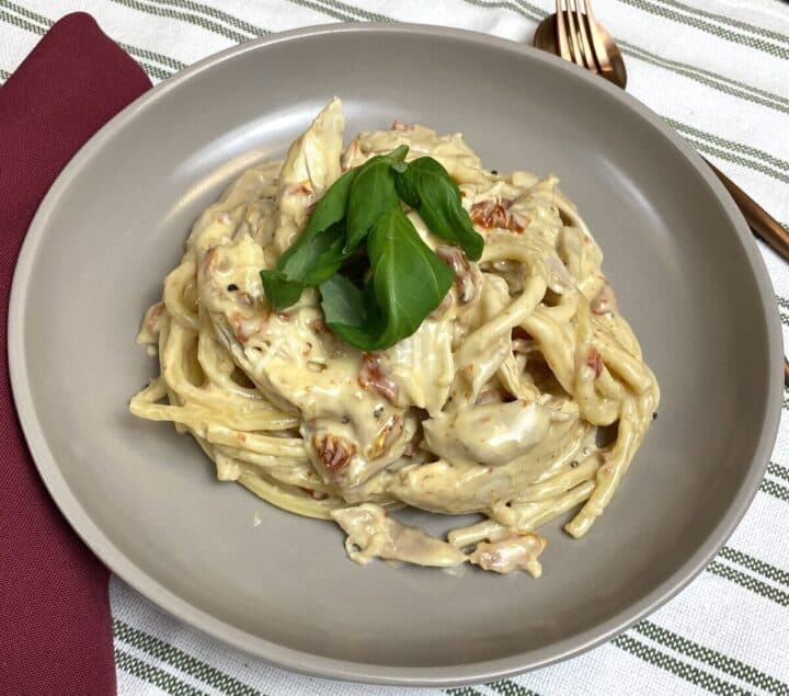 A bowl of marry me chicken pasta topped with fresh basil alongside silverware.