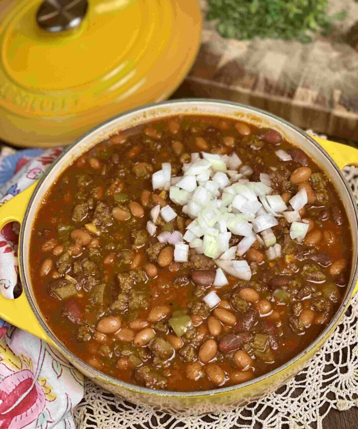 A yellow pot of chili with onion topping alongside the lid and a cutting board.