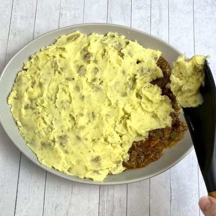 A dish of shepherds pie with a spatula adding more mashed potatoes.