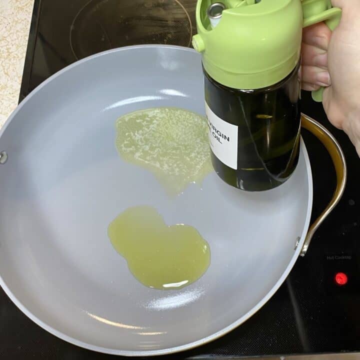 A skillet on the stove with a hand holding a bottle of oil.