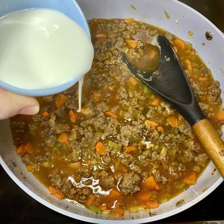 A skillet on the stove with ground meat filling cooking and a hand adding a slurry.