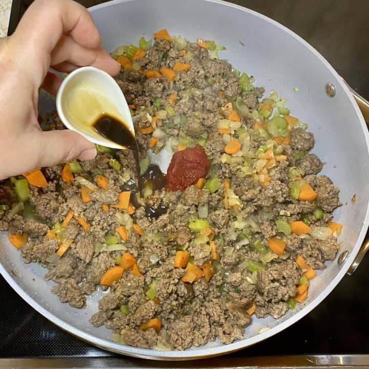 A skillet on the stove with cooked vegetables and meat with a hand adding tomato paste.