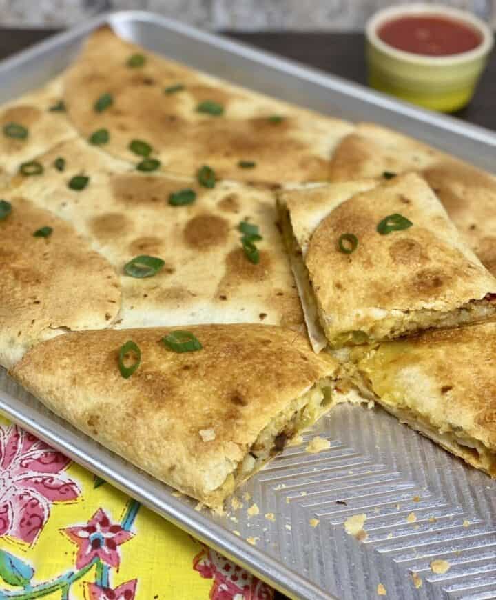 A fully cooked sheet pan quesadilla de papa with a piece cut out and green onions on top.