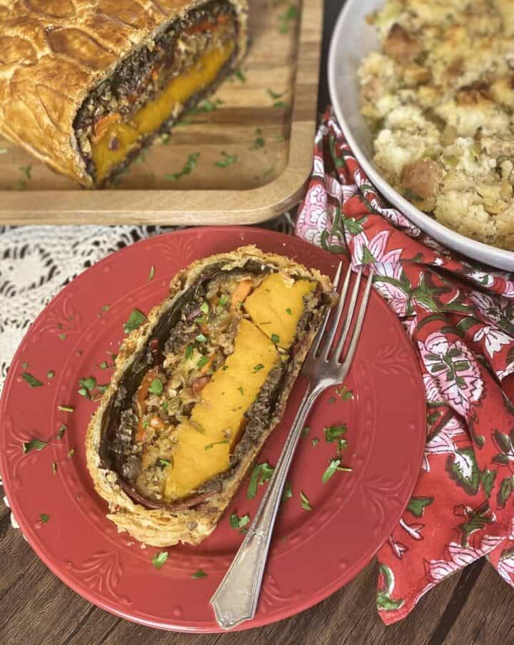 A vegetable wellington loaf on a wood platter with a slice on a plate and a side of stuffing.